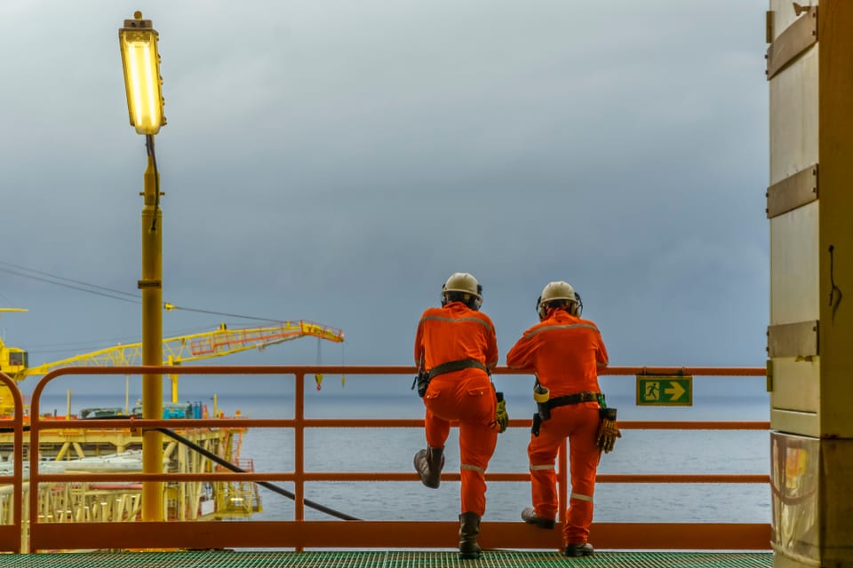 Two offshore workers in orange coveralls and hard hats at a platform railing looking out over a grey sea
