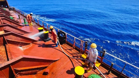 Crew members hosing down the red deck of a cargo ship at sea under clear skies