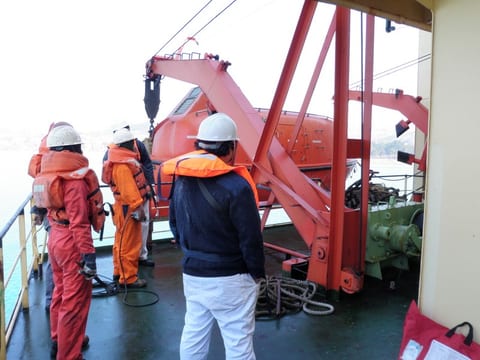 Inspectors in hard hats and lifejackets observing a davit launched enclosed lifeboat during a shipboard drill